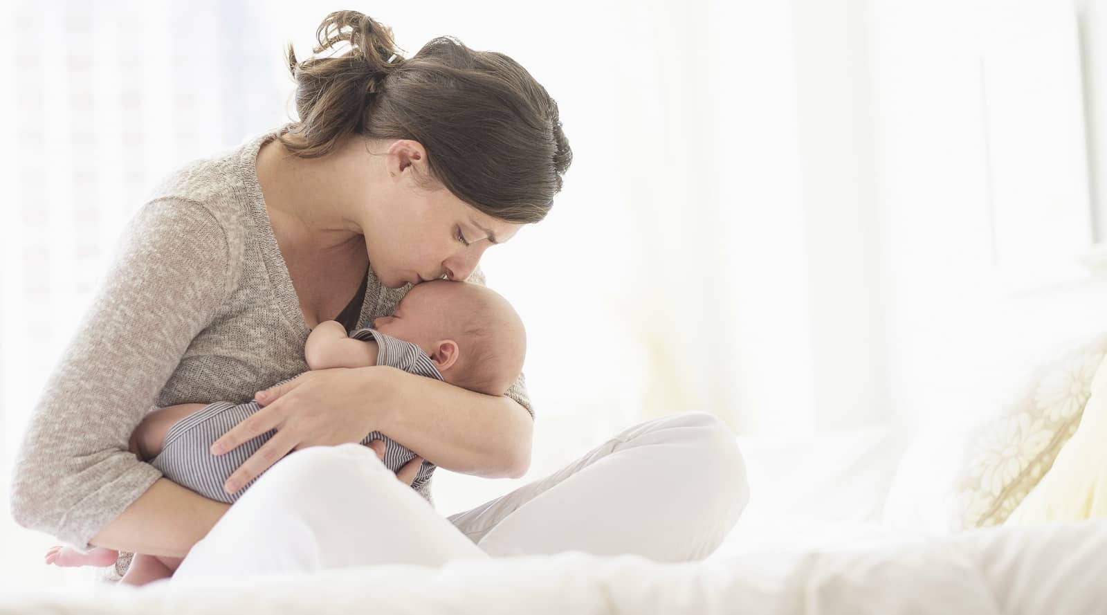 woman siting crossed legged while comforting and kissing a newborn on the forehead