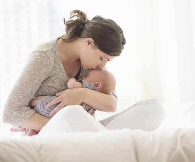 woman siting crossed legged while comforting and kissing a newborn on the forehead