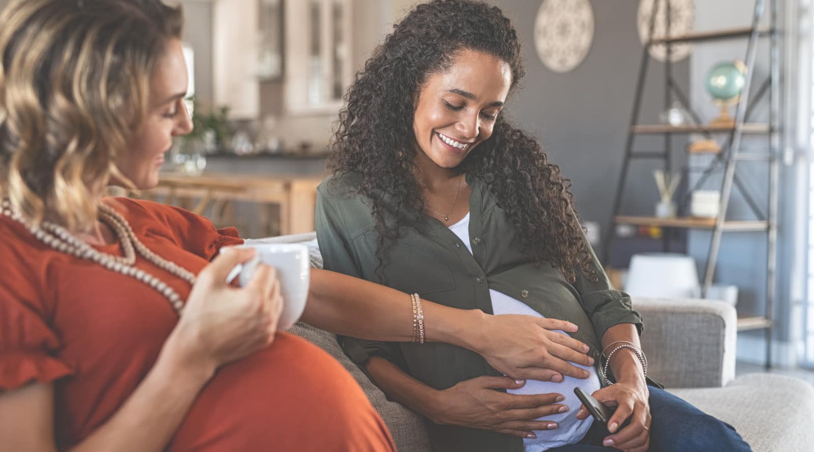 Two pregnant women socialising over a hot beverage. One mother is holding her baby bump and another mother holds her hand out